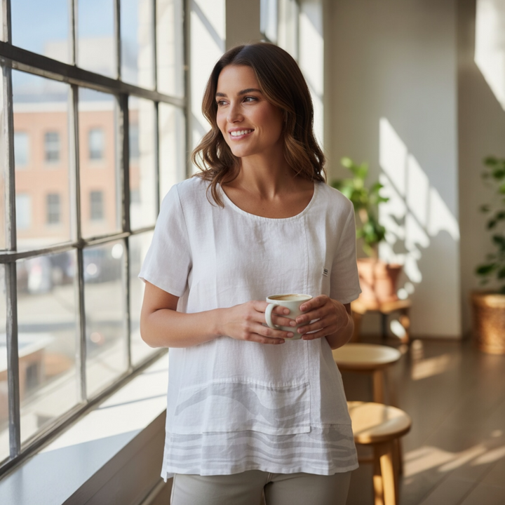 Lady wearing White Sands Linen Top holding a mug in a sunlit room with large windows
