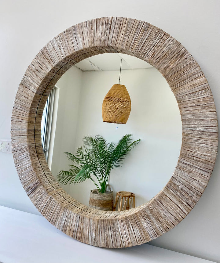 A round mirror with a white wash water hyacinth frame, hanging on a wall above a white surface, with a potted plant and a decorative basket reflected in the mirror.