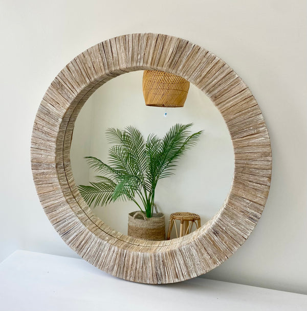 A round mirror with a beige water hyacinth frame, hanging on a wall above a white surface, with a potted plant and a decorative basket reflected in the mirror.