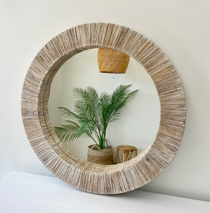 A round mirror with a beige water hyacinth frame, hanging on a wall above a white surface, with a potted plant and a decorative basket reflected in the mirror.