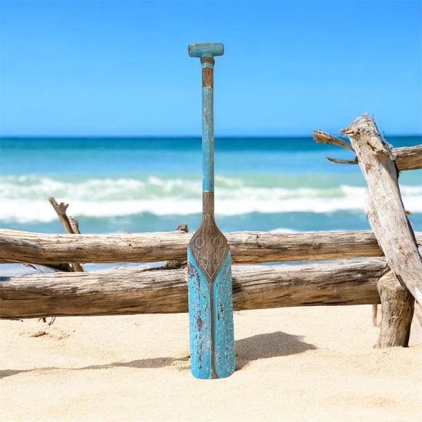 Wooden oar with blue finish leaning against driftwood on a sandy beach with ocean in the background