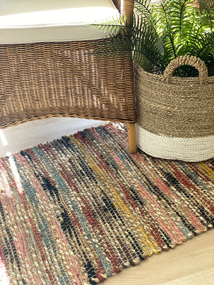 Multicolored braided rug on a wooden floor with a wicker basket and plant in the background.