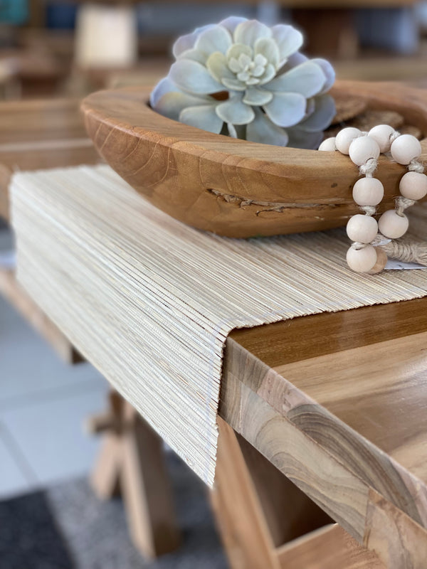 A beige table runner made of natural fibers displayed on a wooden dining table, with a wooden bowl and decorative items in the background.