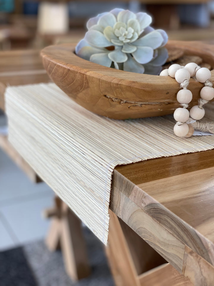 A beige table runner made of natural fibers displayed on a wooden dining table, with a wooden bowl and decorative items in the background.
