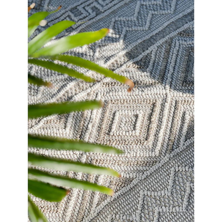 Textured outdoor rug with geometric pattern and green plant leaves.