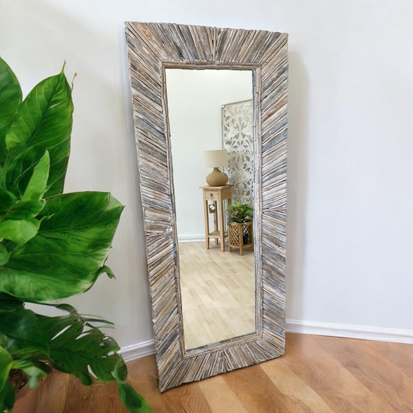 A rectangular driftwood mirror with a white washed frame, placed vertical on a wall, reflecting a part of a room and a plant.