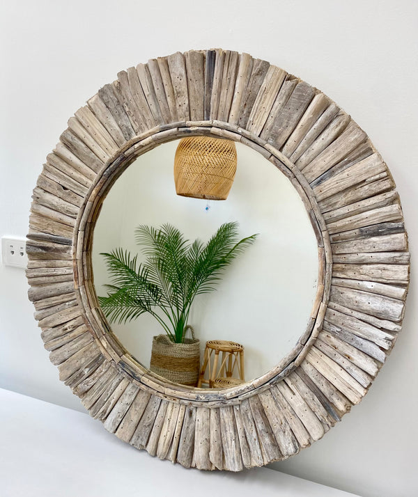 A round driftwood mirror with a white washed frame, hanging on a wall with a reflection of a plant and a wicker basket.