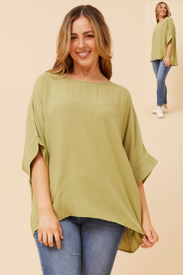 Woman wearing a green loose top over a blue top and jeans on a beige background