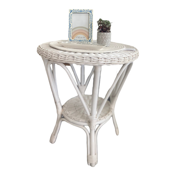 A white, round, cane side table with a lower shelf, displaying a decorative frame and a small potted plant on top.
