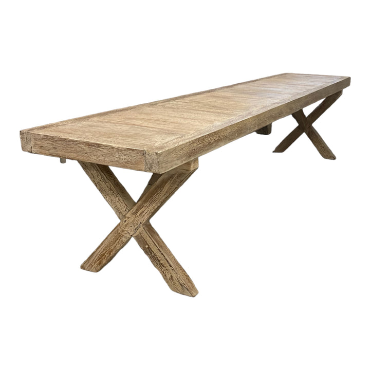 A long, handcrafted wooden bench with a white wash finish and a crossed-leg design, showcasing various wood tones.