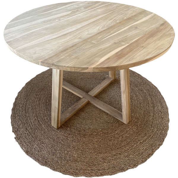 A round teak dining table with square legs placed on a round jute rug. The table is unsealed, showcasing a natural teak wood finish.