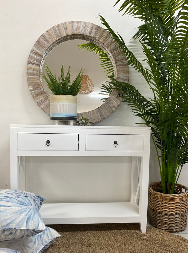 A white console table with drawers, placed against a wall with a large circular mirror above it. The console is flanked by two potted plants, and there are decorative items on the console and pillows on the floor.