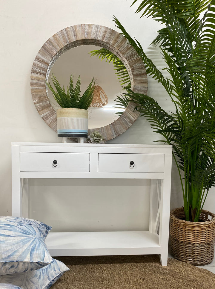 A white console table with drawers, placed against a wall with a large circular mirror above it. The console is flanked by two potted plants, and there are decorative items on the console and pillows on the floor.